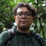 Portrait of a hiker surrounded by dense green jungle, wearing glasses and a green hoodie.